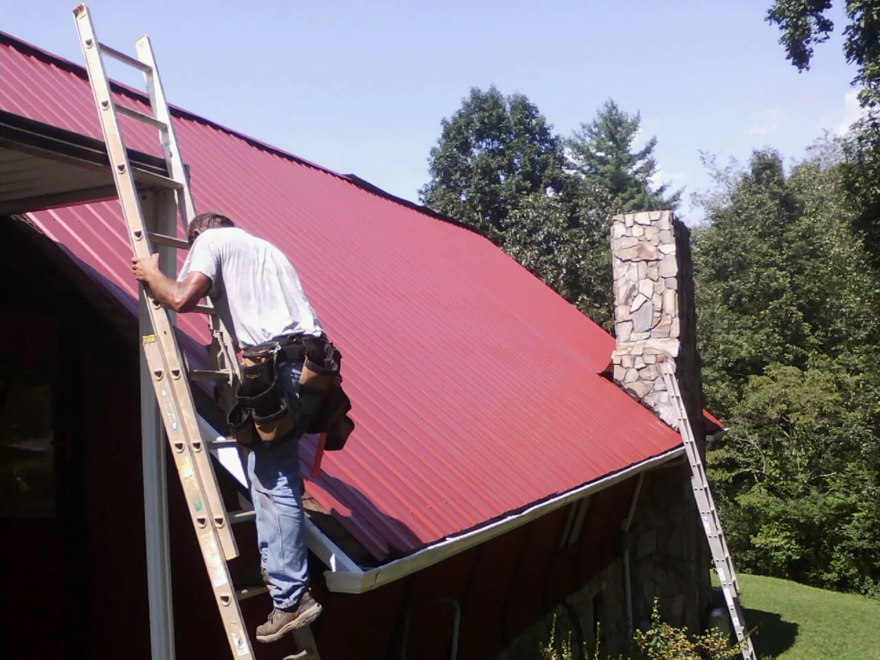 Roofer on a ladder beside a red metal roof and stone chimney — Soffit & Fascia Repair by Roofing Brothers in Apple Valley